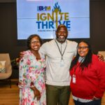 Photo of three attendees smile and pose together in front of a screen displaying the 2025 BHM Small Business Week 'Ignite and Thrive: Fueling Small Business' event branding