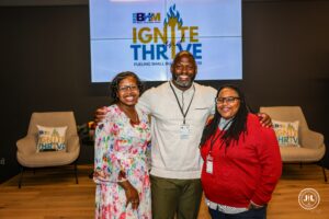 Photo of three attendees smile and pose together in front of a screen displaying the 2025 BHM Small Business Week 'Ignite and Thrive: Fueling Small Business' event branding