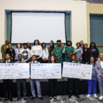 Photo of a large group of youth and adults pose together holding oversized checks for 1st place ($750), 2nd place ($500), and 3rd place ($250) at the Small Business Week 2025 Youth Pitch competition.