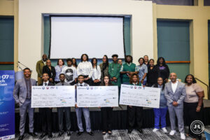 Photo of a large group of youth and adults pose together holding oversized checks for 1st place ($750), 2nd place ($500), and 3rd place ($250) at the Small Business Week 2025 Youth Pitch competition.