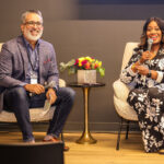 Two speakers seated in armchairs on a stage, each holding a microphone, smiling during a panel discussion. A small table with a floral arrangement sits between them.