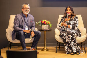 Two speakers seated in armchairs on a stage, each holding a microphone, smiling during a panel discussion. A small table with a floral arrangement sits between them.