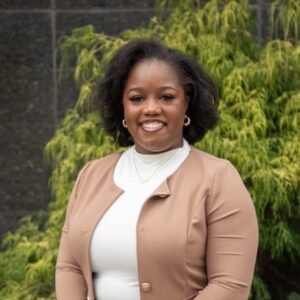 Photo headshot of Reinvest Plan Officer, Archie Stewart. African American woman with short hair wearing a white shirt and tan blazer.