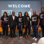 Photo of six people posed together in front of a 'Welcome' screen at a Black Men Buy Houses event, with several wearing branded black t-shirts.