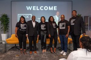 Photo of six people posed together in front of a 'Welcome' screen at a Black Men Buy Houses event, with several wearing branded black t-shirts.