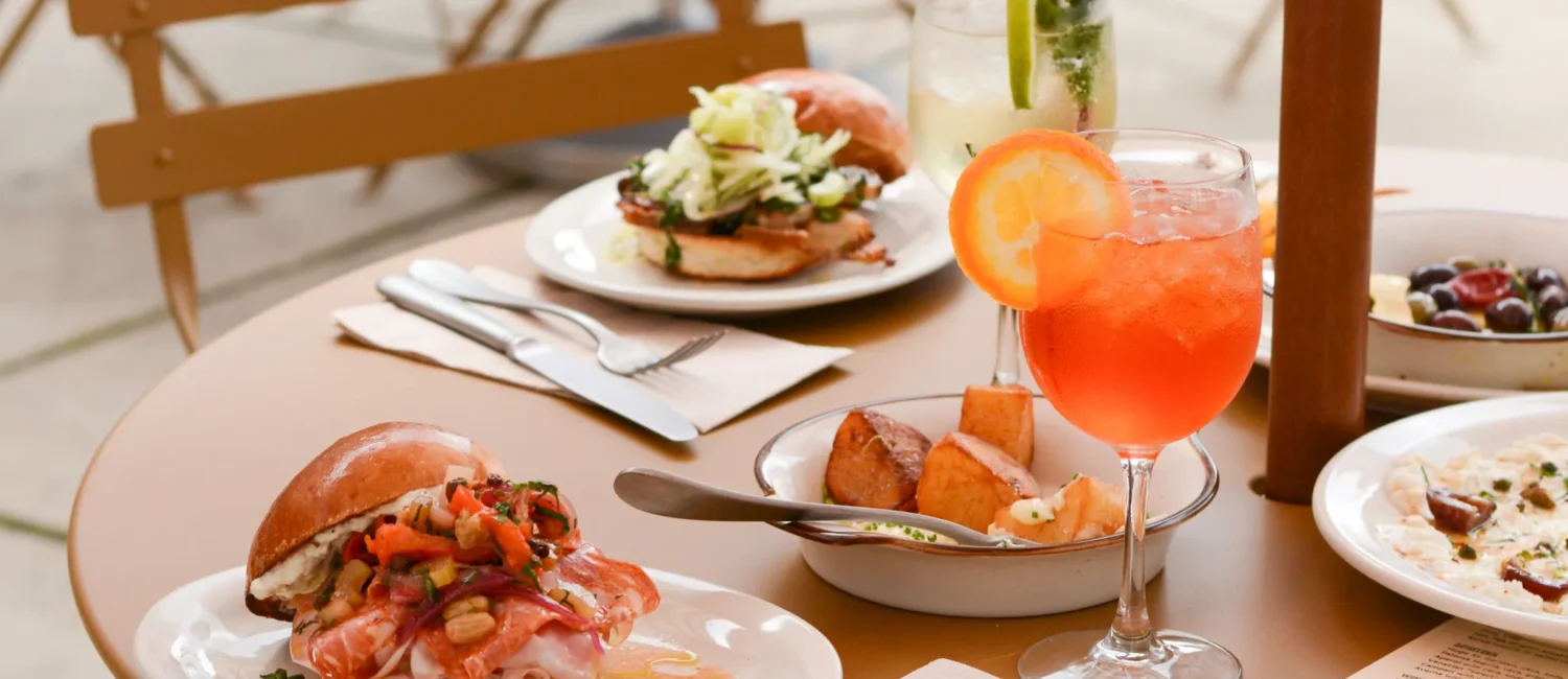 A close-up of a round, tan outdoor cafe table set with a variety of dishes and drinks. In the foreground is a brioche sandwich loaded with thinly sliced cured meats and a chunky relish, sitting next to a bright orange spritz cocktail garnished with an orange slice. The table is also filled with roasted potatoes, another sandwich topped with shredded greens, a clear cocktail with mint and lime, and various small appetizer plates.