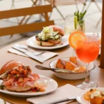 A close-up of a round, tan outdoor cafe table set with a variety of dishes and drinks. In the foreground is a brioche sandwich loaded with thinly sliced cured meats and a chunky relish, sitting next to a bright orange spritz cocktail garnished with an orange slice. The table is also filled with roasted potatoes, another sandwich topped with shredded greens, a clear cocktail with mint and lime, and various small appetizer plates.
