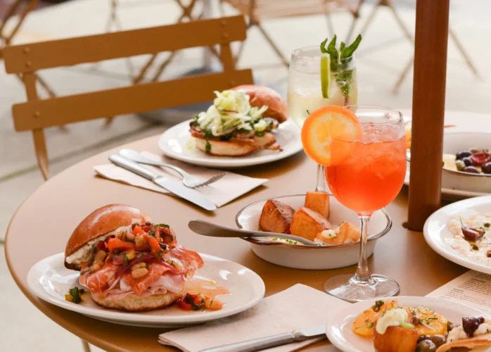 A close-up of a round, tan outdoor cafe table set with a variety of dishes and drinks. In the foreground is a brioche sandwich loaded with thinly sliced cured meats and a chunky relish, sitting next to a bright orange spritz cocktail garnished with an orange slice. The table is also filled with roasted potatoes, another sandwich topped with shredded greens, a clear cocktail with mint and lime, and various small appetizer plates.