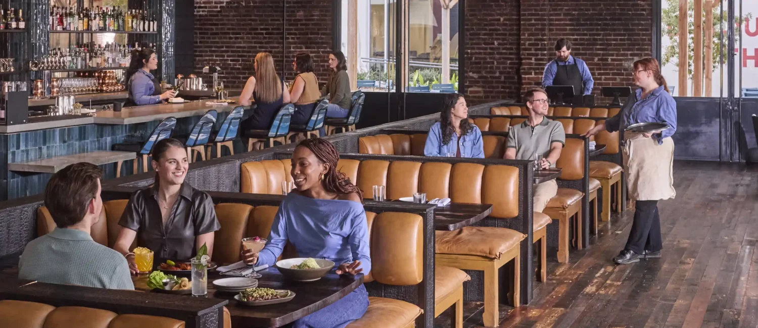 People dining in a bustling, modern restaurant with exposed brick walls and hardwood floors. In the foreground, three friends smile and chat over food and cocktails in a tan leather booth. In the background, other patrons sit at similar booths and at a blue-tiled bar, while staff members attend to the guests.