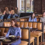People dining in a bustling, modern restaurant with exposed brick walls and hardwood floors. In the foreground, three friends smile and chat over food and cocktails in a tan leather booth. In the background, other patrons sit at similar booths and at a blue-tiled bar, while staff members attend to the guests.