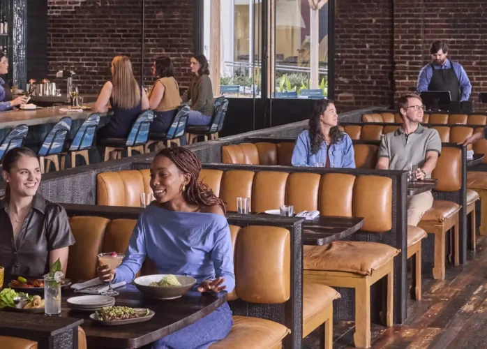 People dining in a bustling, modern restaurant with exposed brick walls and hardwood floors. In the foreground, three friends smile and chat over food and cocktails in a tan leather booth. In the background, other patrons sit at similar booths and at a blue-tiled bar, while staff members attend to the guests.