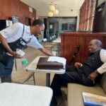A barista wearing a black apron smiling warmly as he serves a beverage to a cheerful older gentleman seated at a booth inside a cafe with wood-paneled walls.