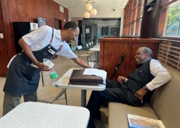 A barista wearing a black apron smiling warmly as he serves a beverage to a cheerful older gentleman seated at a booth inside a cafe with wood-paneled walls.