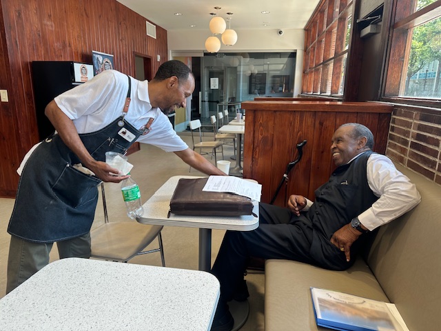 A barista wearing a black apron smiling warmly as he serves a beverage to a cheerful older gentleman seated at a booth inside a cafe with wood-paneled walls.