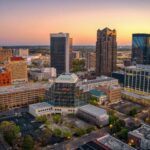 An aerial view of The City of Birmingham skyline at sunset, featuring a mix of modern high-rises and historic brick buildings surrounded by lush green trees.