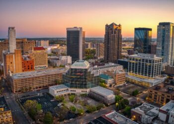 An aerial view of The City of Birmingham skyline at sunset, featuring a mix of modern high-rises and historic brick buildings surrounded by lush green trees.