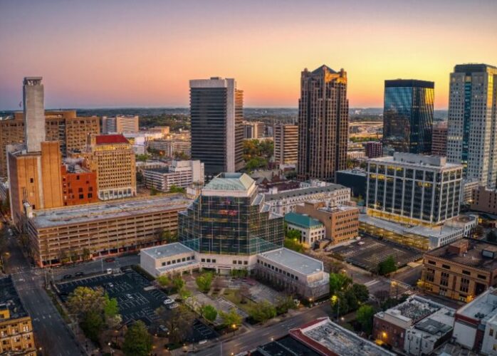 An aerial view of The City of Birmingham skyline at sunset, featuring a mix of modern high-rises and historic brick buildings surrounded by lush green trees.