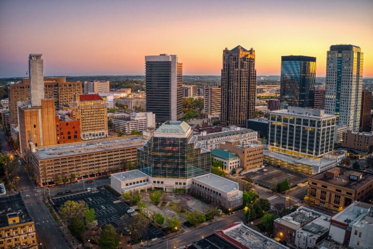 An aerial view of The City of Birmingham skyline at sunset, featuring a mix of modern high-rises and historic brick buildings surrounded by lush green trees.