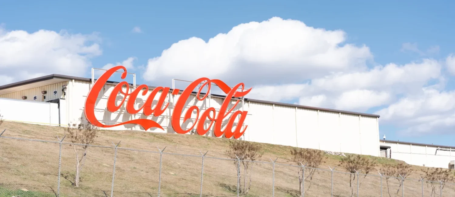 : A large, classic red Coca-Cola sign mounted on the side of a long white industrial building that sits atop a grassy hill against a blue sky with fluffy clouds.