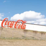 : A large, classic red Coca-Cola sign mounted on the side of a long white industrial building that sits atop a grassy hill against a blue sky with fluffy clouds.