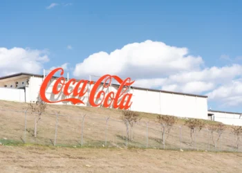 : A large, classic red Coca-Cola sign mounted on the side of a long white industrial building that sits atop a grassy hill against a blue sky with fluffy clouds.