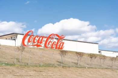 : A large, classic red Coca-Cola sign mounted on the side of a long white industrial building that sits atop a grassy hill against a blue sky with fluffy clouds.