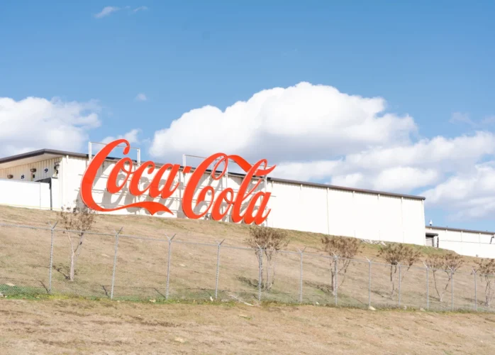 : A large, classic red Coca-Cola sign mounted on the side of a long white industrial building that sits atop a grassy hill against a blue sky with fluffy clouds.