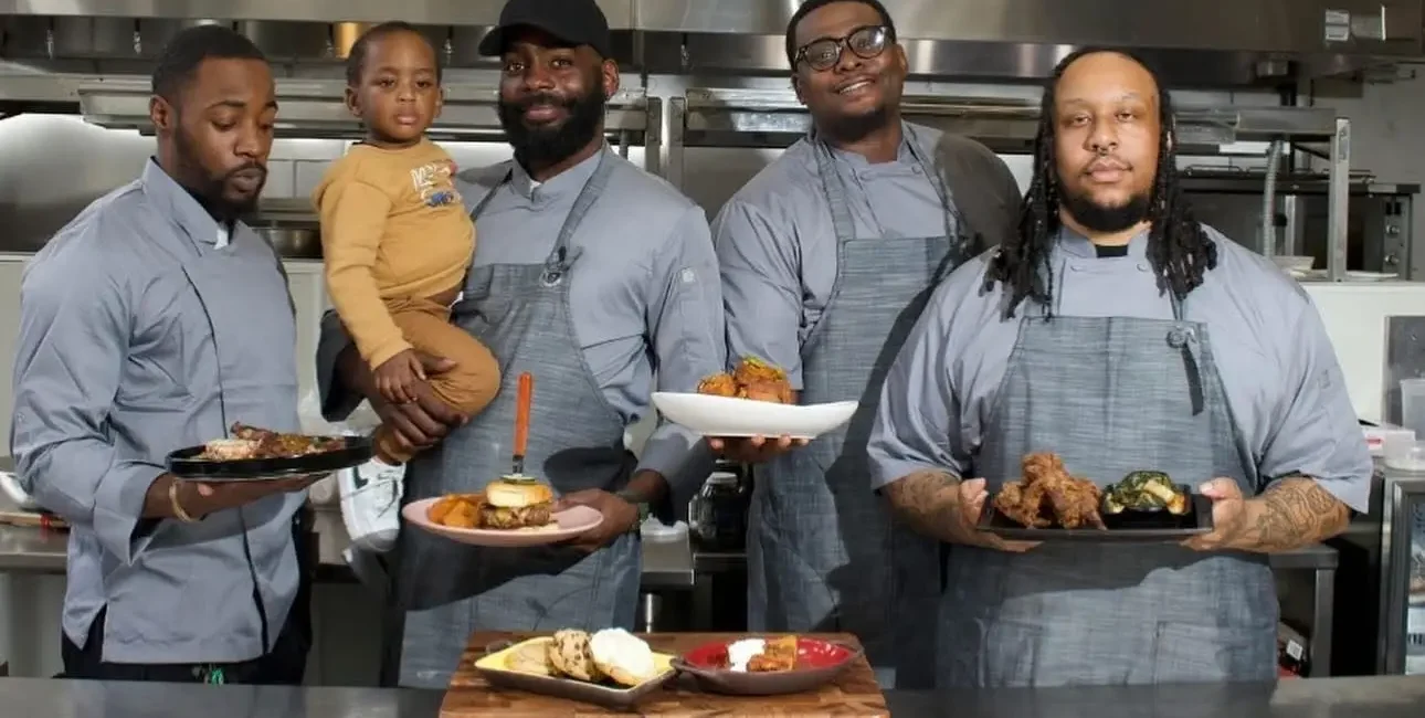 Four male chefs in matching grey aprons standing side-by-side in a commercial kitchen, proudly holding up plates of food including a burger and fried chicken. One chef is holding a young toddler.