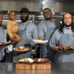 Four male chefs in matching grey aprons standing side-by-side in a commercial kitchen, proudly holding up plates of food including a burger and fried chicken. One chef is holding a young toddler.