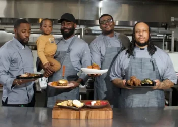 Four male chefs in matching grey aprons standing side-by-side in a commercial kitchen, proudly holding up plates of food including a burger and fried chicken. One chef is holding a young toddler.