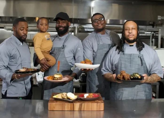 Four male chefs in matching grey aprons standing side-by-side in a commercial kitchen, proudly holding up plates of food including a burger and fried chicken. One chef is holding a young toddler.