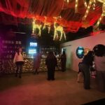 The interior of a dimly lit, festive bar decorated with red fabric draping and illuminated wire reindeer hanging from the ceiling. Patrons mingle in groups near a gold-patterned bar counter and next to a bright pink neon sign on the left wall.