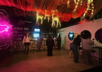 The interior of a dimly lit, festive bar decorated with red fabric draping and illuminated wire reindeer hanging from the ceiling. Patrons mingle in groups near a gold-patterned bar counter and next to a bright pink neon sign on the left wall.