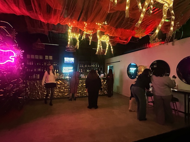 The interior of a dimly lit, festive bar decorated with red fabric draping and illuminated wire reindeer hanging from the ceiling. Patrons mingle in groups near a gold-patterned bar counter and next to a bright pink neon sign on the left wall.