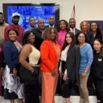 A large, diverse group of smiling professionals posing together in a brightly lit room, positioned next to a blue graphic with the "Small Business Intensive" logo.
