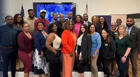 A large, diverse group of smiling professionals posing together in a brightly lit room, positioned next to a blue graphic with the "Small Business Intensive" logo.