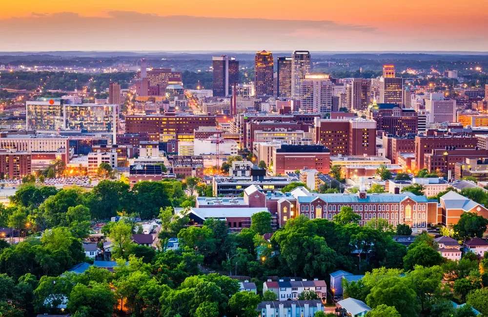 An aerial view of The City of Birmingham skyline at sunset, featuring a mix of modern high-rises and historic brick buildings surrounded by lush green trees.