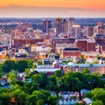 An aerial view of The City of Birmingham skyline at sunset, featuring a mix of modern high-rises and historic brick buildings surrounded by lush green trees.
