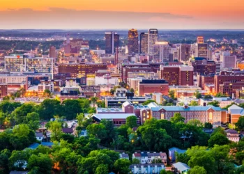 An aerial view of The City of Birmingham skyline at sunset, featuring a mix of modern high-rises and historic brick buildings surrounded by lush green trees.
