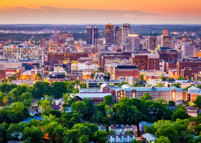 An aerial view of The City of Birmingham skyline at sunset, featuring a mix of modern high-rises and historic brick buildings surrounded by lush green trees.