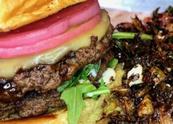A close-up shot of a juicy double cheeseburger topped with bright pink pickled red onions and arugula on a toasted bun, served next to a side of crispy roasted Brussels sprouts with cheese crumbles