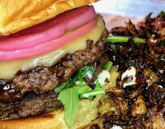 A close-up shot of a juicy double cheeseburger topped with bright pink pickled red onions and arugula on a toasted bun, served next to a side of crispy roasted Brussels sprouts with cheese crumbles