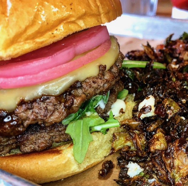A close-up shot of a juicy double cheeseburger topped with bright pink pickled red onions and arugula on a toasted bun, served next to a side of crispy roasted Brussels sprouts with cheese crumbles