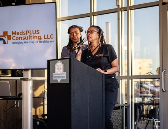 : A woman in dark scrubs speaking into a microphone at a Negro Southern League Museum podium. A screen to her left displays the logo for MedsPLUS Consulting, LLC.