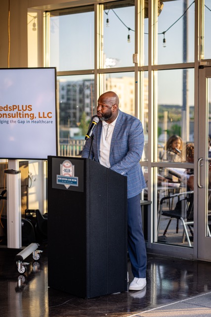 A man wearing a blue plaid jacket and white shirt speaking at a Negro Southern League Museum podium next to a digital display showing the MedsPLUS Consulting, LLC logo.