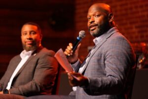 A man in a blue plaid blazer speaking into a microphone while seated on a stage during a panel or presentation, with another man in a dark suit sitting beside him listening.