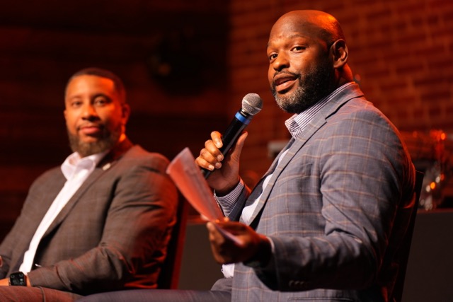 A man in a blue plaid blazer speaking into a microphone while seated on a stage during a panel or presentation, with another man in a dark suit sitting beside him listening.