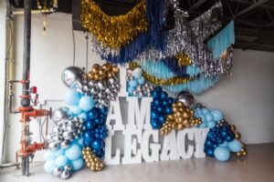 A festive event photo backdrop featuring large white block letters spelling "I AM LEGACY," surrounded by a large arch of light blue, dark blue, gold, and silver balloons, with metallic fringe hanging from the ceiling above.