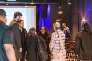 A candid photo of a diverse group of attendees mingling at an indoor event, centered on a woman with glasses and long dark curly hair laughing heartily.