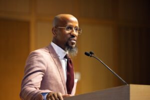 A man with a bald head, glasses, and a long goatee speaking at a podium. He is dressed in a mauve blazer, white collared shirt, and dark tie.